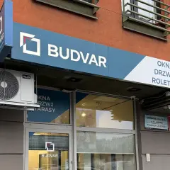 Logo firmy BUDVAR z napisem "OKNA DRZWI ROLETY" na tle pomarańczowej ściany.