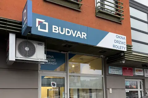 Logo firmy BUDVAR z napisem "OKNA DRZWI ROLETY" na tle pomarańczowej ściany.